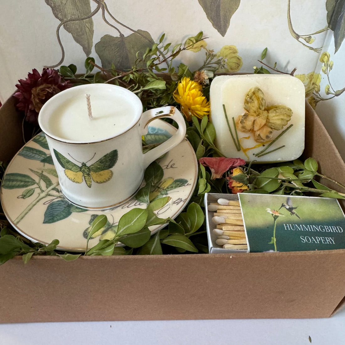 kraft gift box with cup and saucer with soy candle inside, all natural soap with soap butterfly on front, surrounded by dried botanicals and flowers